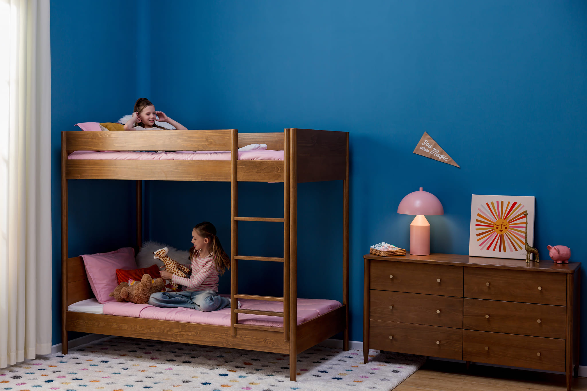 A blue-walled bedroom with a modern bunk bed with pink bed sheets.   A matching dresser can be seen next to the bed. Young girls are on each bunk, one paying with a stuffed toy. 