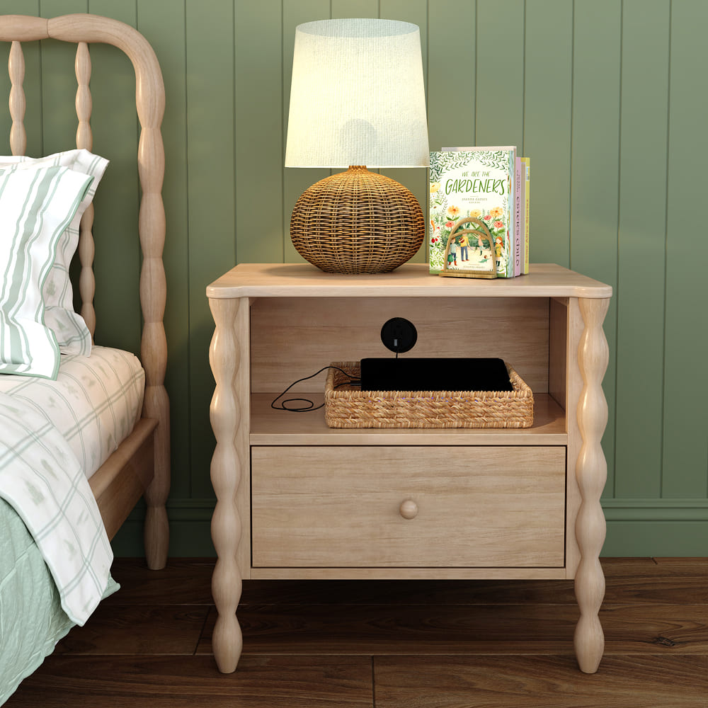 A light-wood finish Jenny Lind style nightstand next to a matching bed in a green room. The nightstand has a shelf with a charger port, holding a basket containing a device. On top of the nightstand is a lamp and book. 