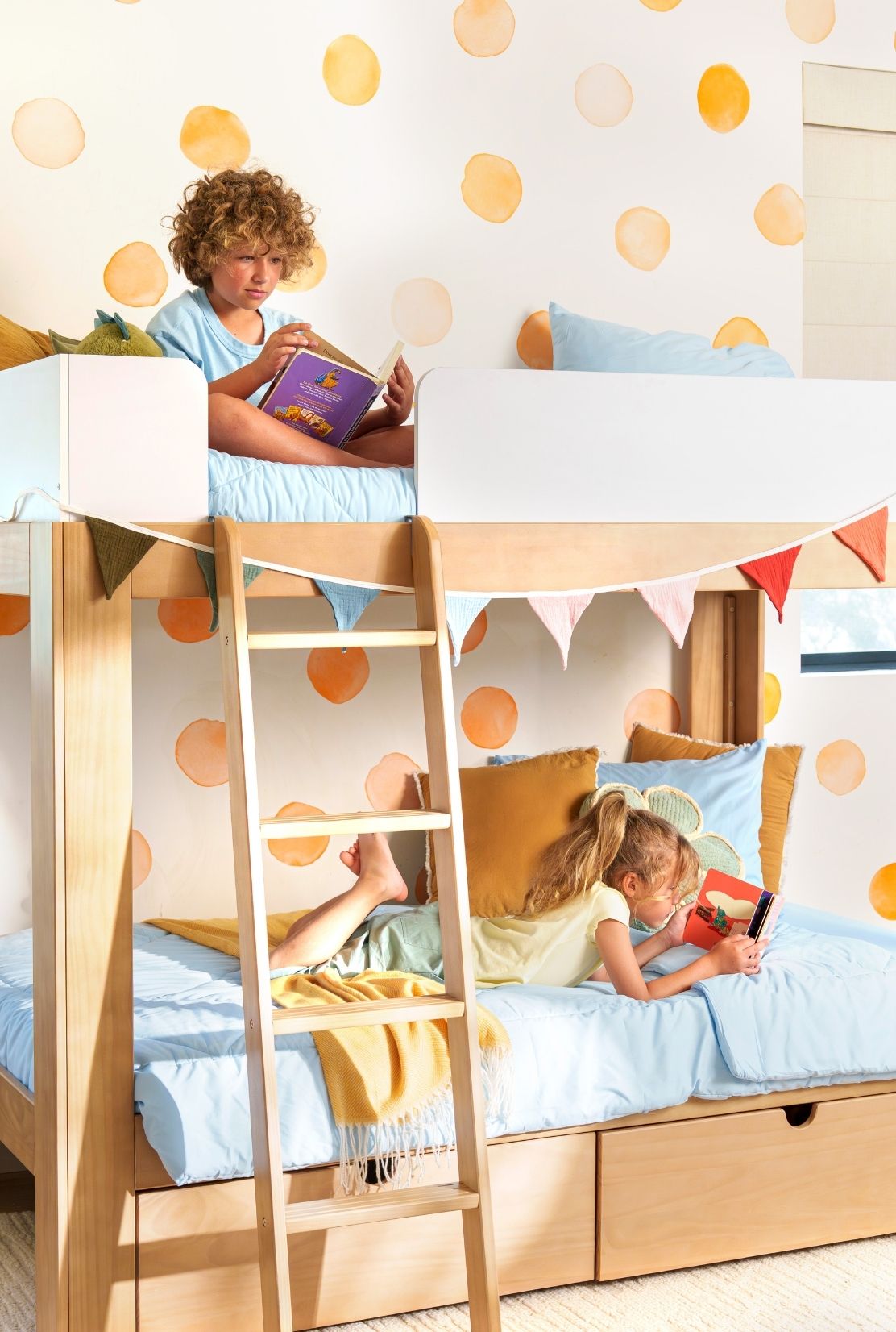 Brother and sister reading in their white and natural wood bunk bed with orange, watercolor polka dot wall decals.