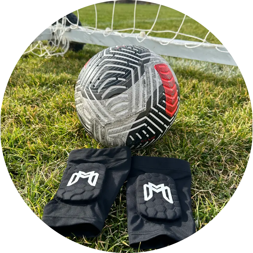 Soccer ball and knee pads on grass near a goal net.