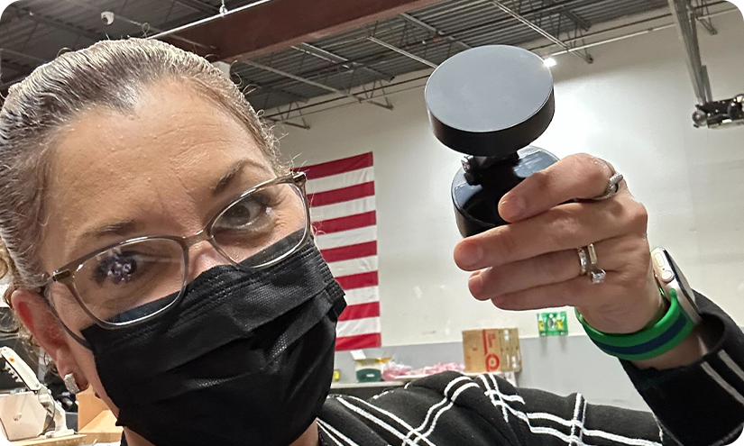 Person wearing a mask holding a small black device, with an American flag in the background.