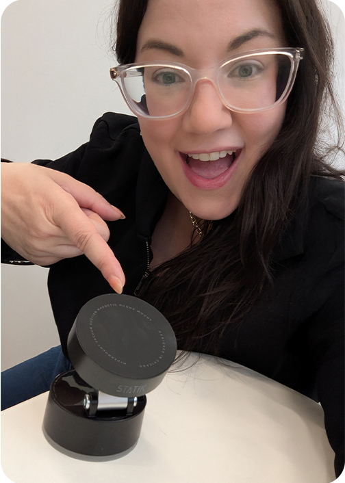 Person smiling and pointing at a round black device on a table.