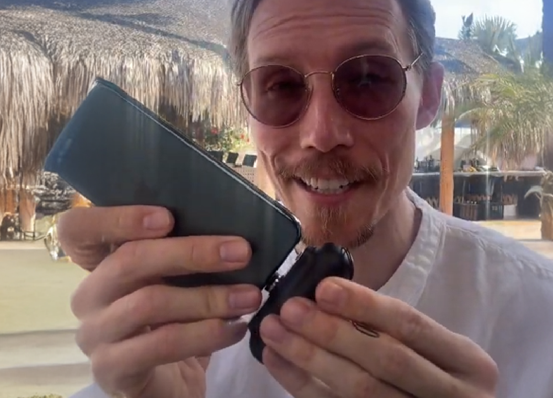 Person holding a smartphone and a small device, smiling outdoors.