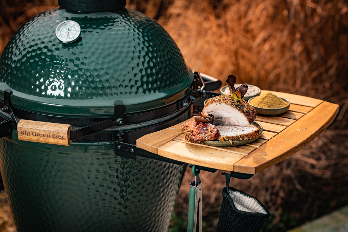 Green ceramic grill with seasoned pork and spices on a wooden side table.