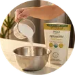 A hand pours a white sweetener from a measuring cup into a metal bowl, with a product bag nearby.