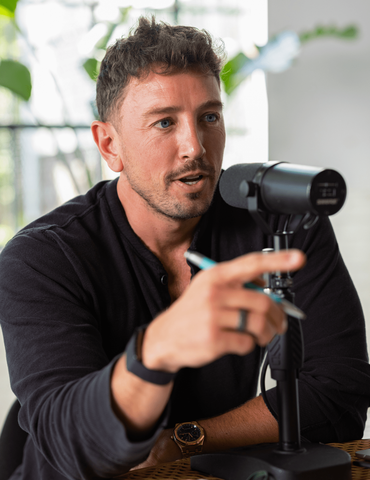 A man in a black shirt speaks into a microphone while gesturing with a pen.