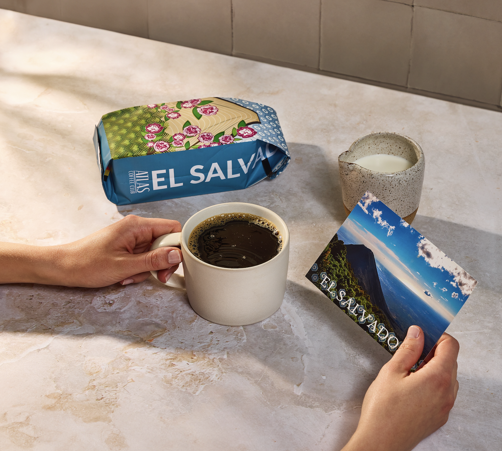 Person holding coffee mug and El Salvador postcard on a table.