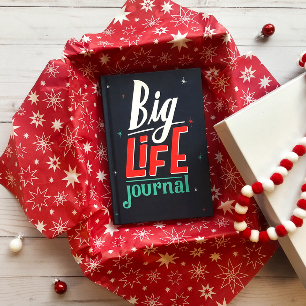 A 'Big Life Journal' sits in a gift box with red, star-patterned tissue paper and ornaments.