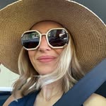 A woman with blonde hair wears a large straw hat, oversized sunglasses, and a seatbelt.