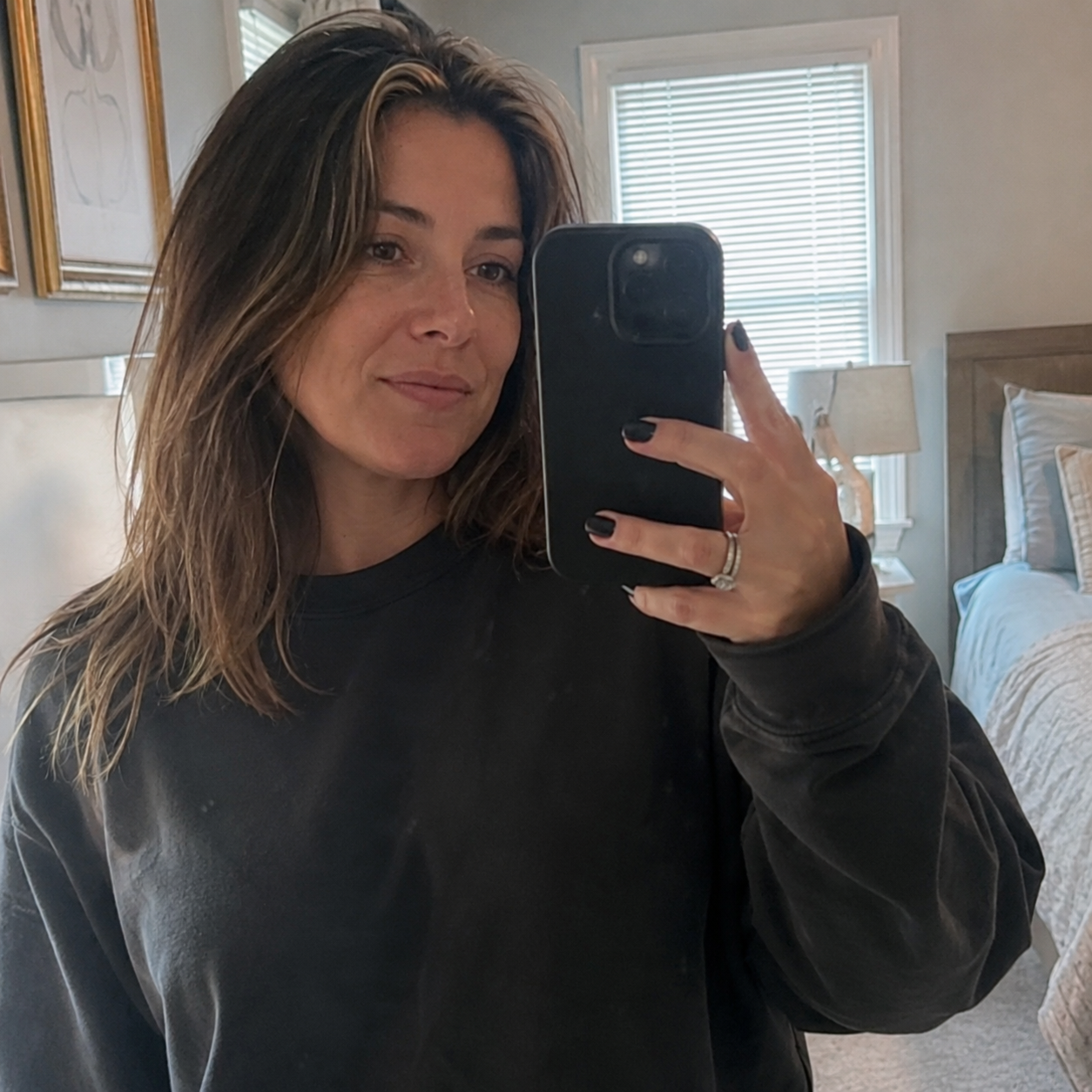 A woman with long brown hair takes a selfie in a mirror inside a bedroom.