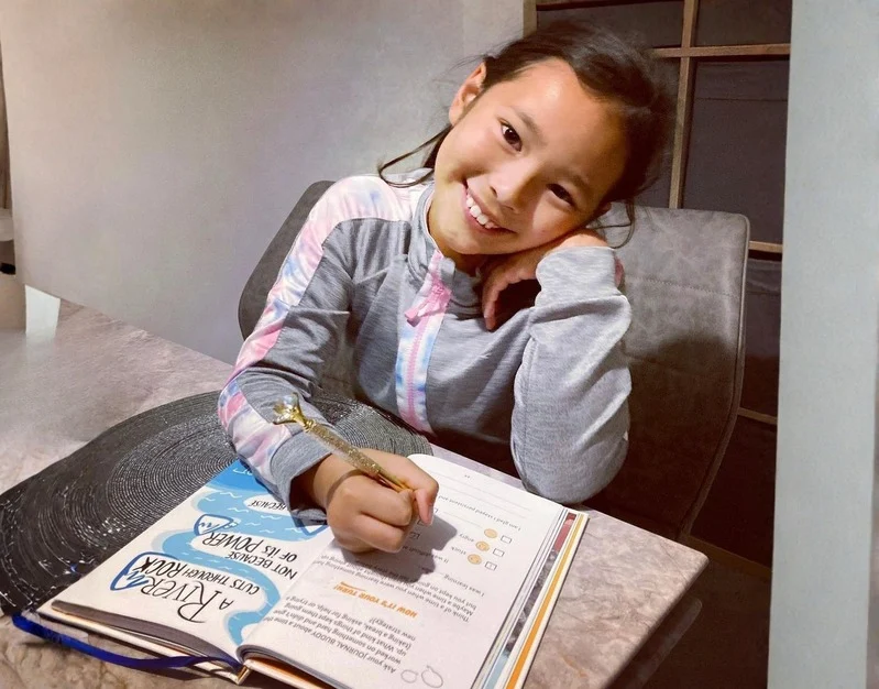 A young girl with dark hair smiles at the camera while writing in a book at a table.