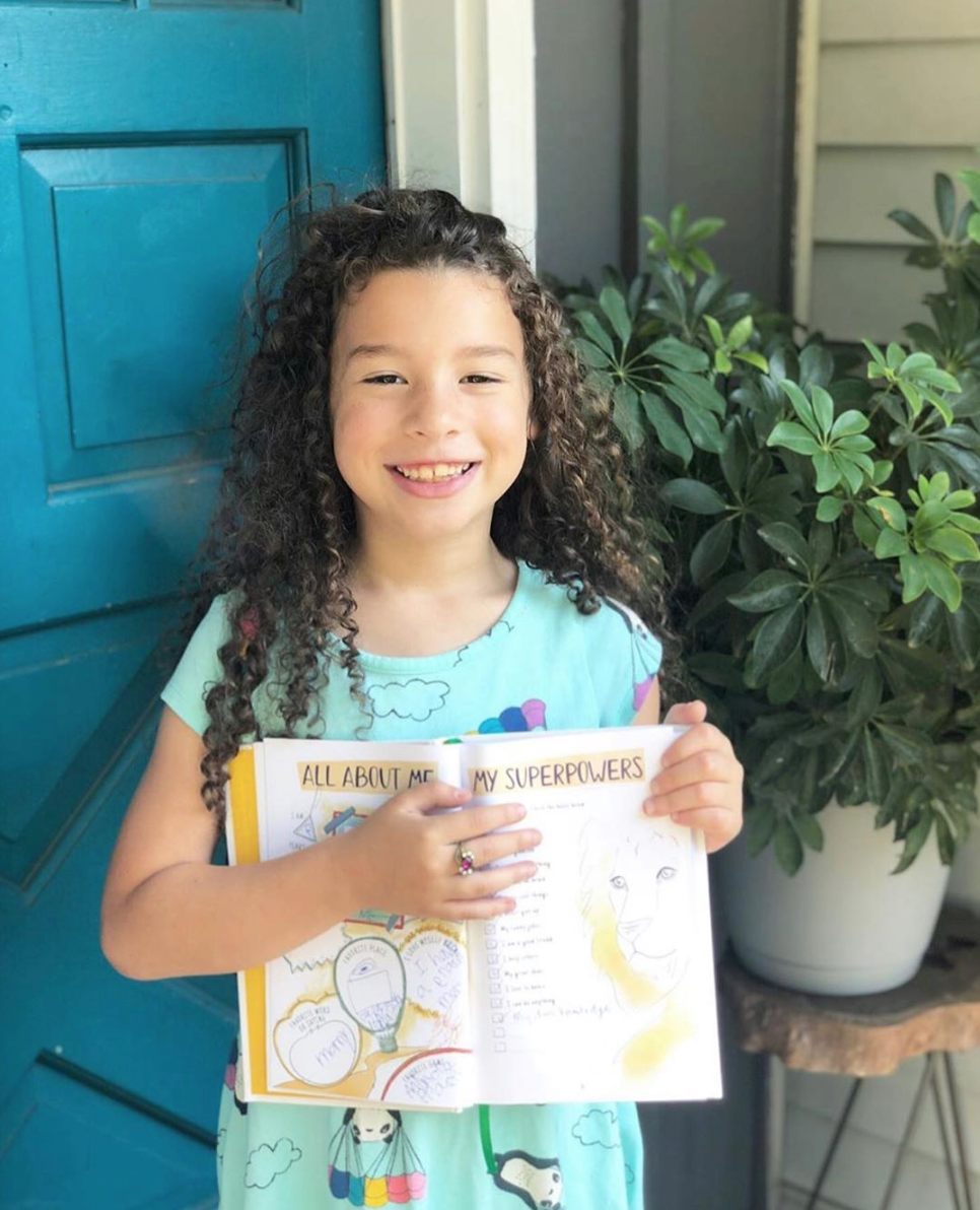A smiling young girl with curly hair holds up an open workbook titled 'All About Me My Superpowers'.