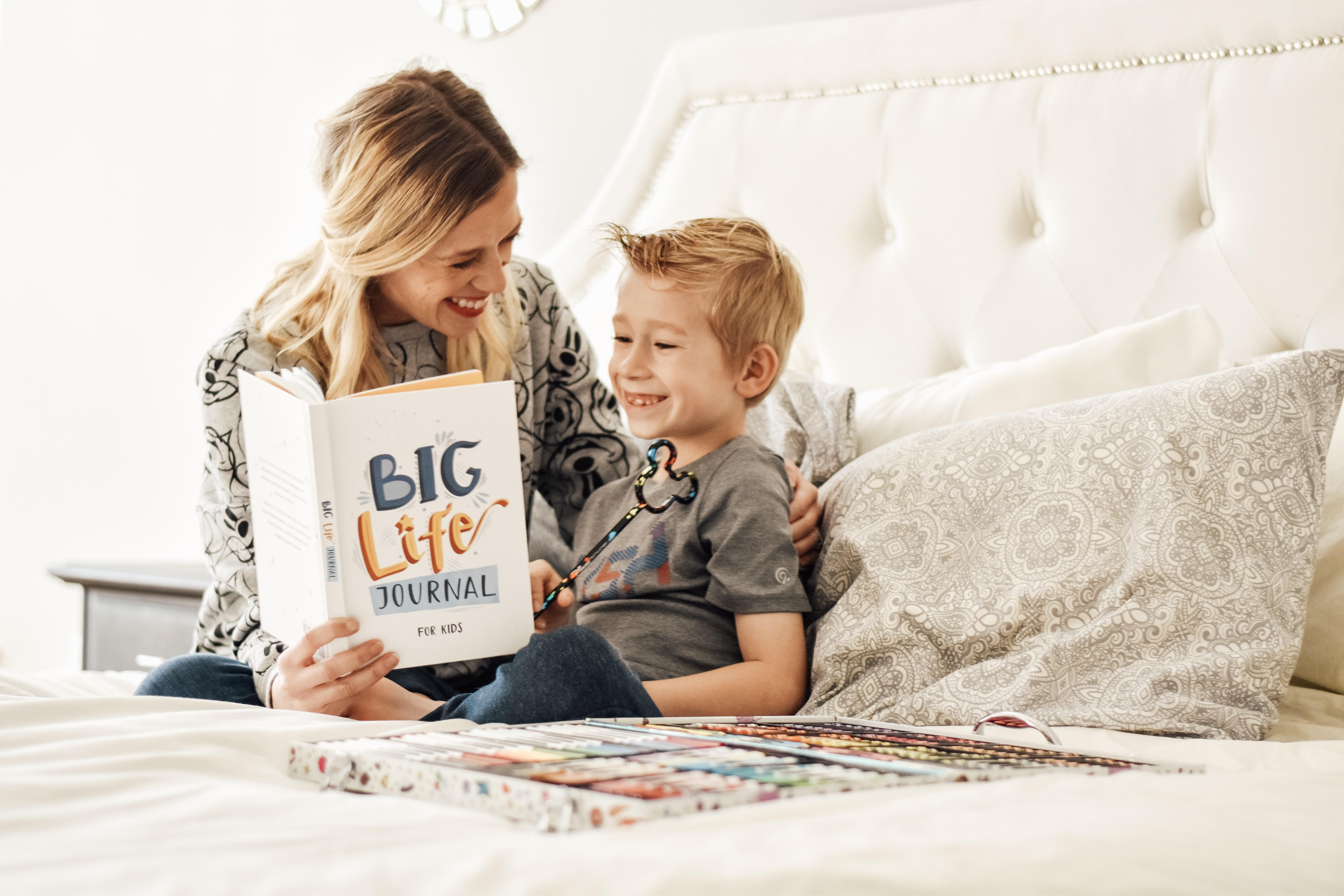 A mother and her young son smile while reading a 'Big Life Journal for Kids' in bed.