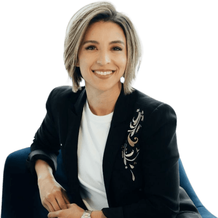 A smiling woman with short hair, wearing a black blazer with embroidery, sitting in a blue chair.