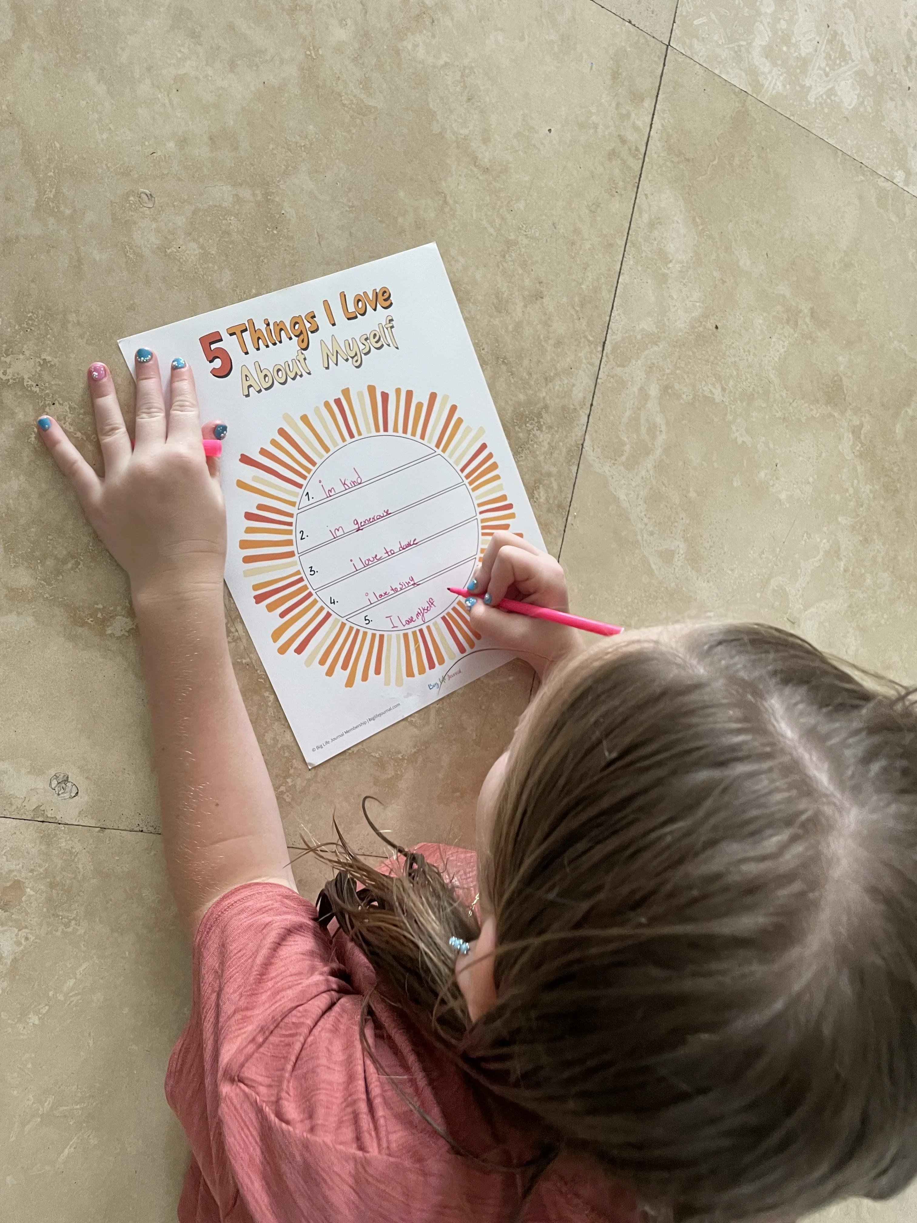 An overhead view of a child filling out a worksheet titled '5 Things I Love About Myself' on the floor.