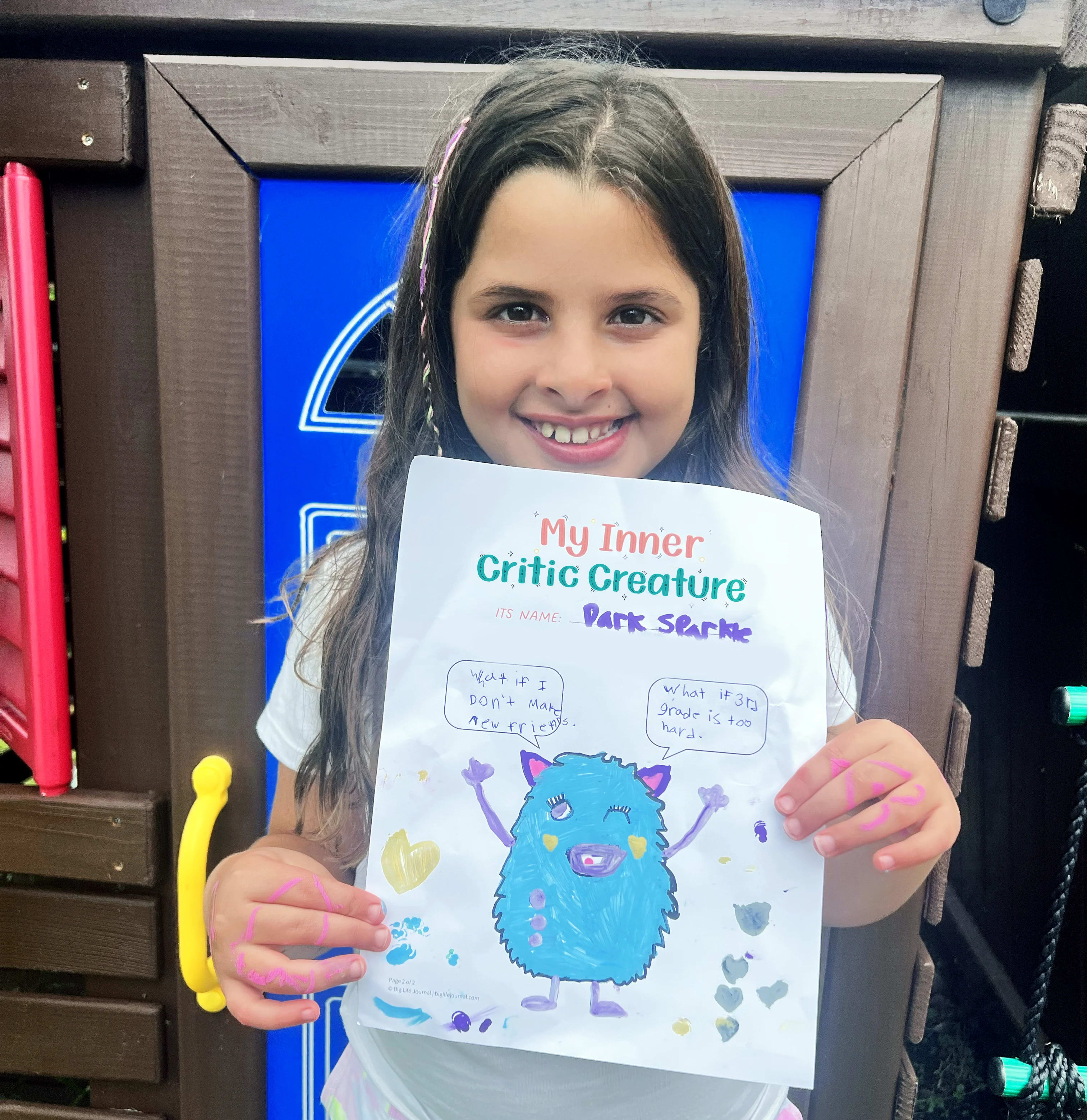 A smiling young girl holds up her drawing of a blue monster titled 'My Inner Critic Creature'.