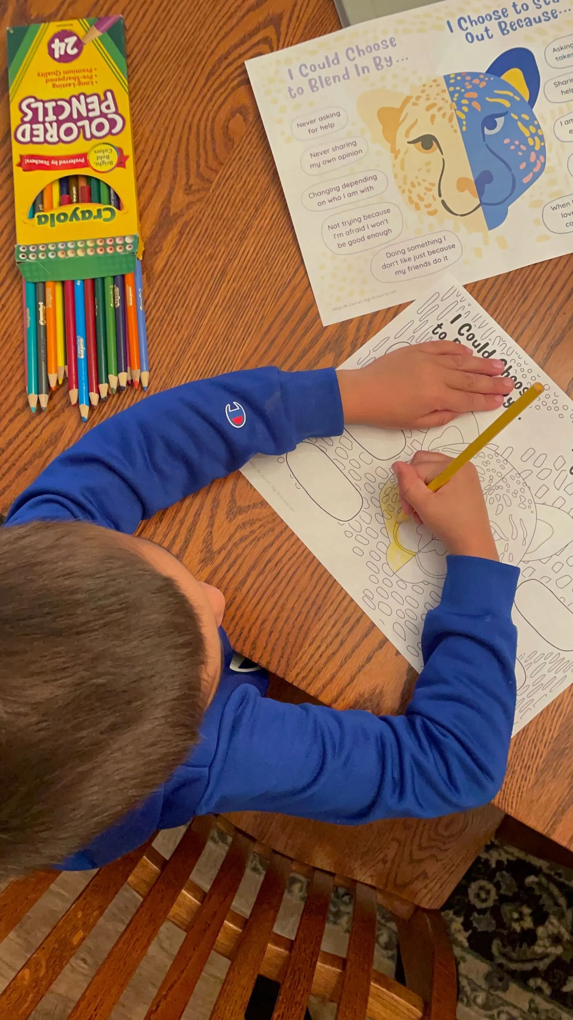An overhead view of a child in a blue shirt coloring a worksheet with a yellow pencil.