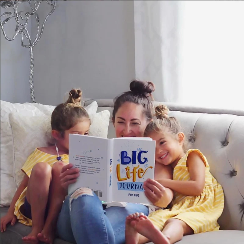 A mother reads the 'Big Life Journal for Kids' to her two smiling daughters on a couch.