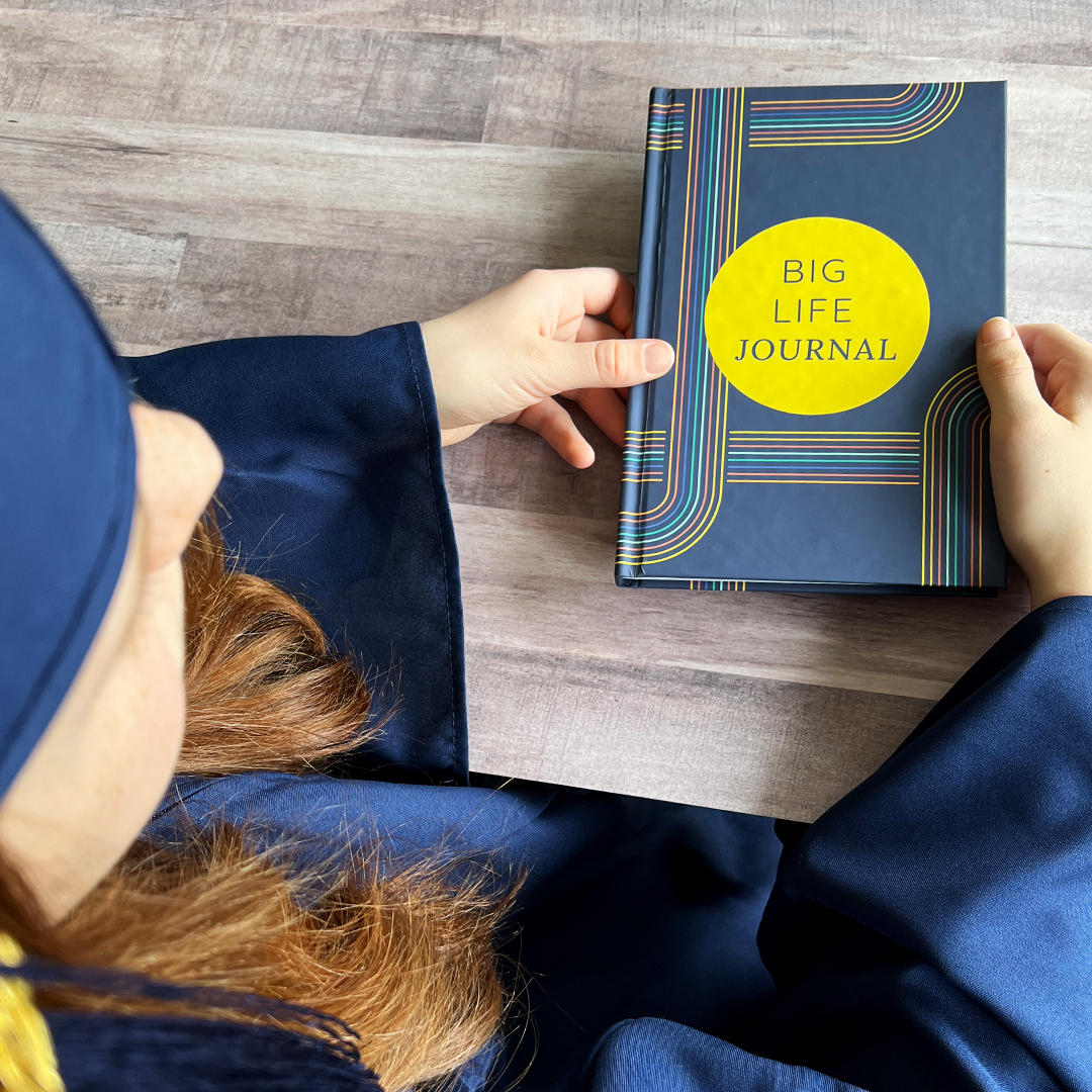 An overhead view of a person in a graduation gown holding a book titled 'Big Life Journal'.