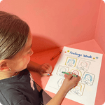 A young girl sits at a desk coloring a worksheet about feelings with a green marker.