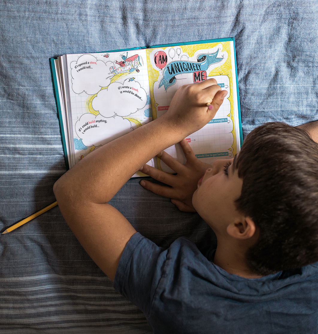 An overhead view of a young boy lying on his stomach on a bed, writing in a journal.