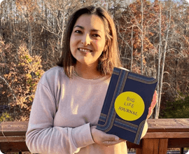 A smiling woman outdoors holds up a blue book titled 'Big Life Journal'.