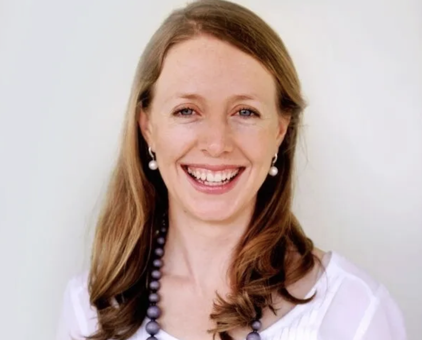 A headshot of a smiling woman with light brown hair, wearing a white shirt and beaded necklace.