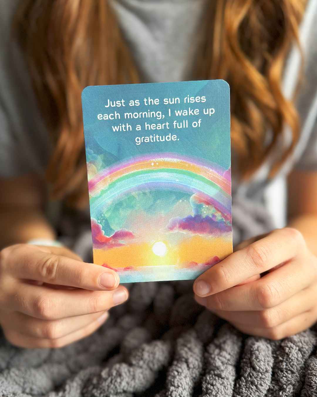 A person holds a gratitude affirmation card featuring a colorful sunrise and a rainbow.