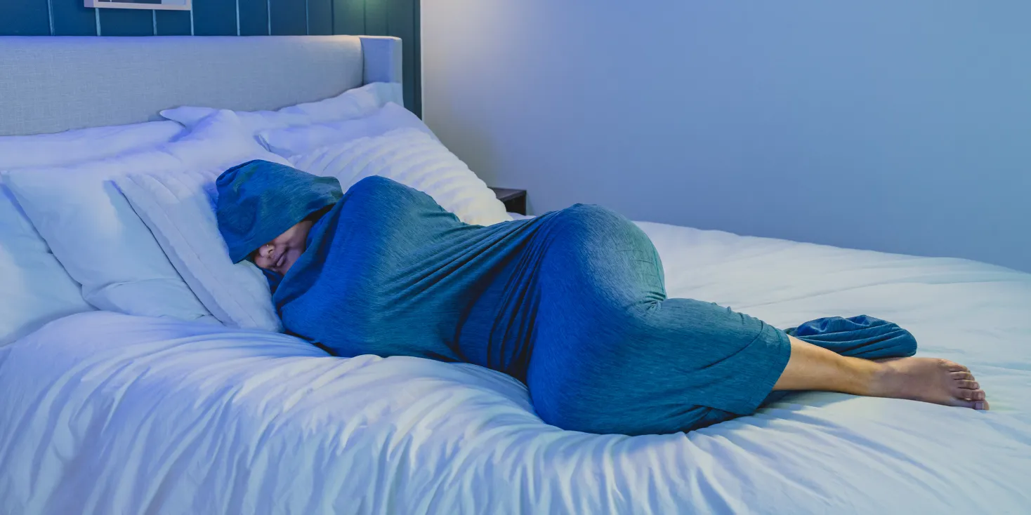 A person lies on their side in bed, fully enclosed in a hooded blue sleep sack.