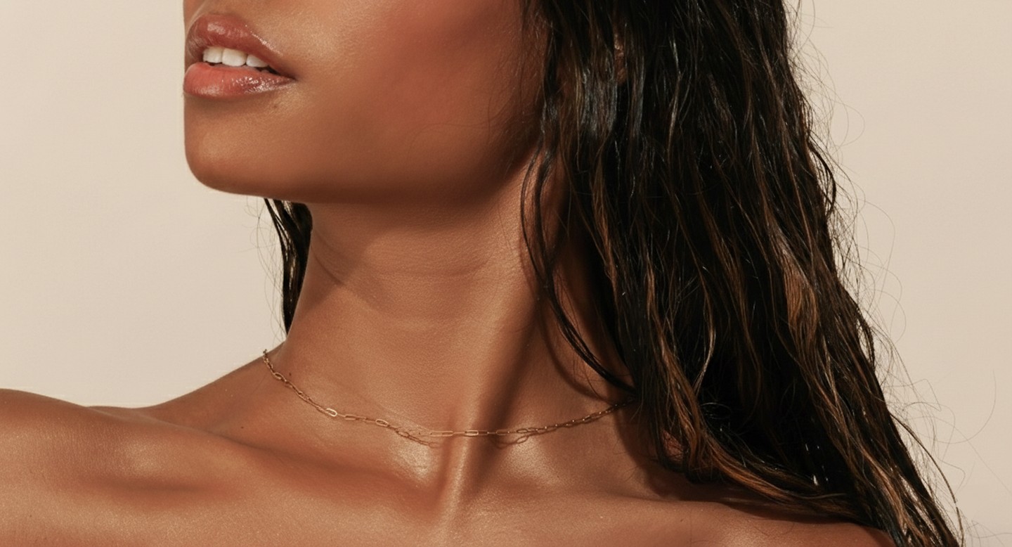 A close-up of a woman with glowing skin wearing a delicate gold chain necklace around her neck.