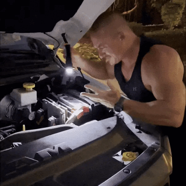 Man in a tank top looking frustrated at an open car hood at night.