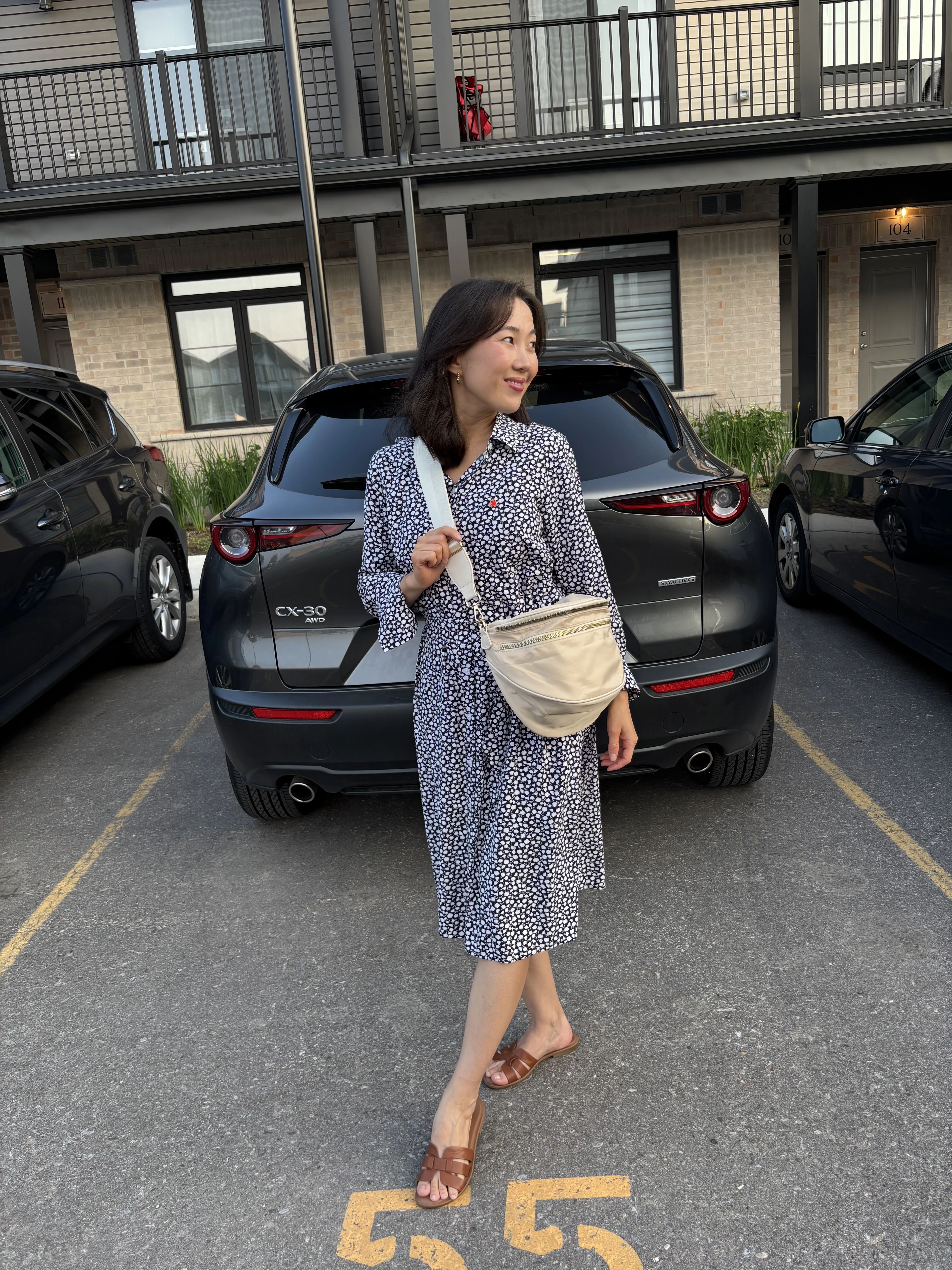 Person standing in front of a dark car in a parking lot, wearing a patterned dress and sandals.