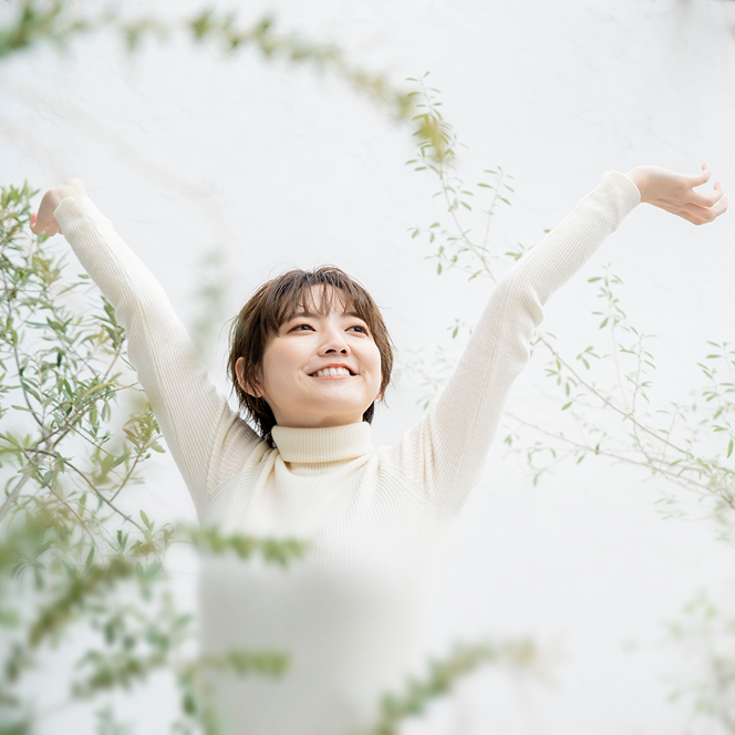 Person in a white sweater smiling with arms raised, surrounded by greenery.