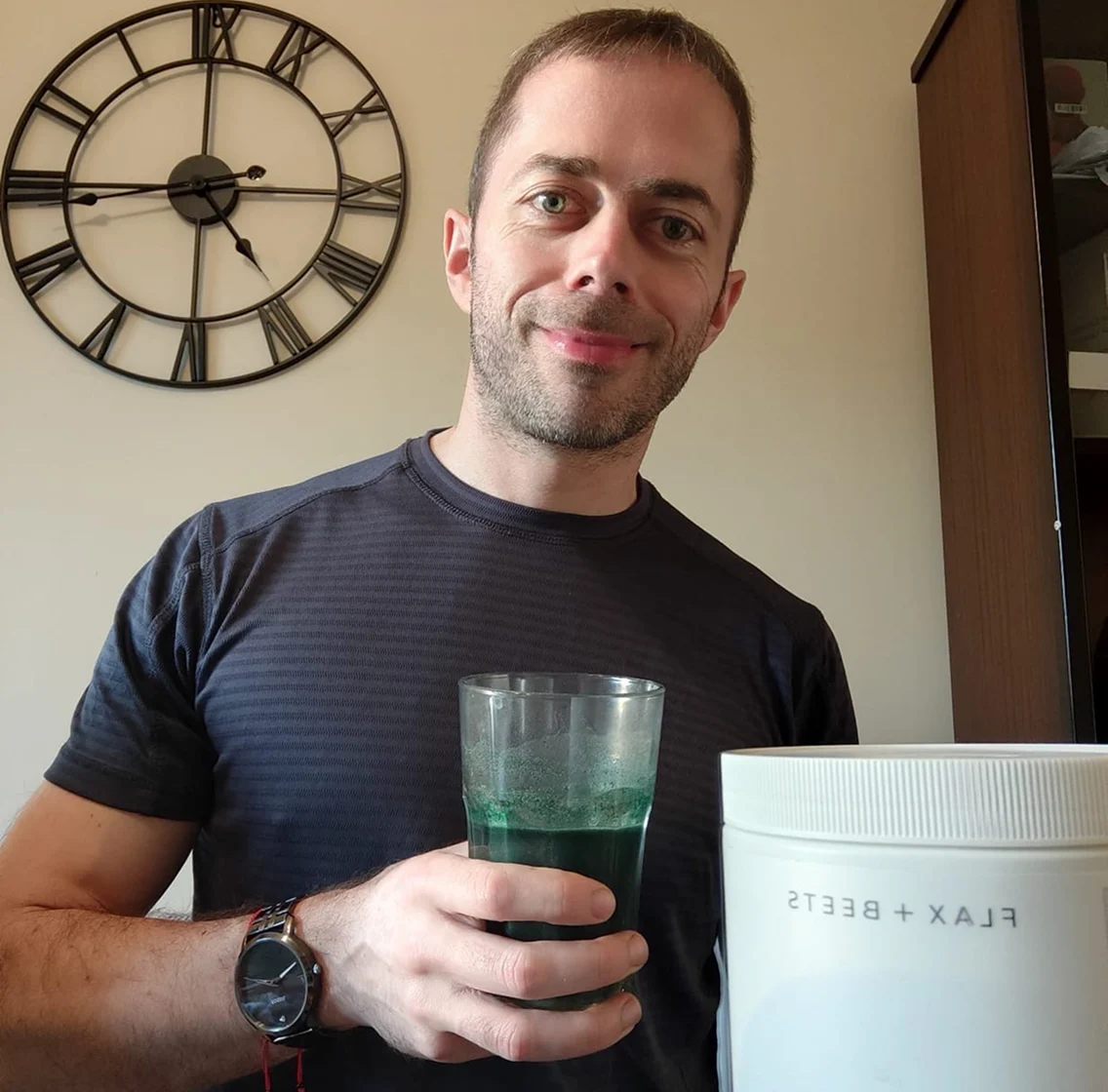 A man smiles while holding a glass of a green drink next to a white container.