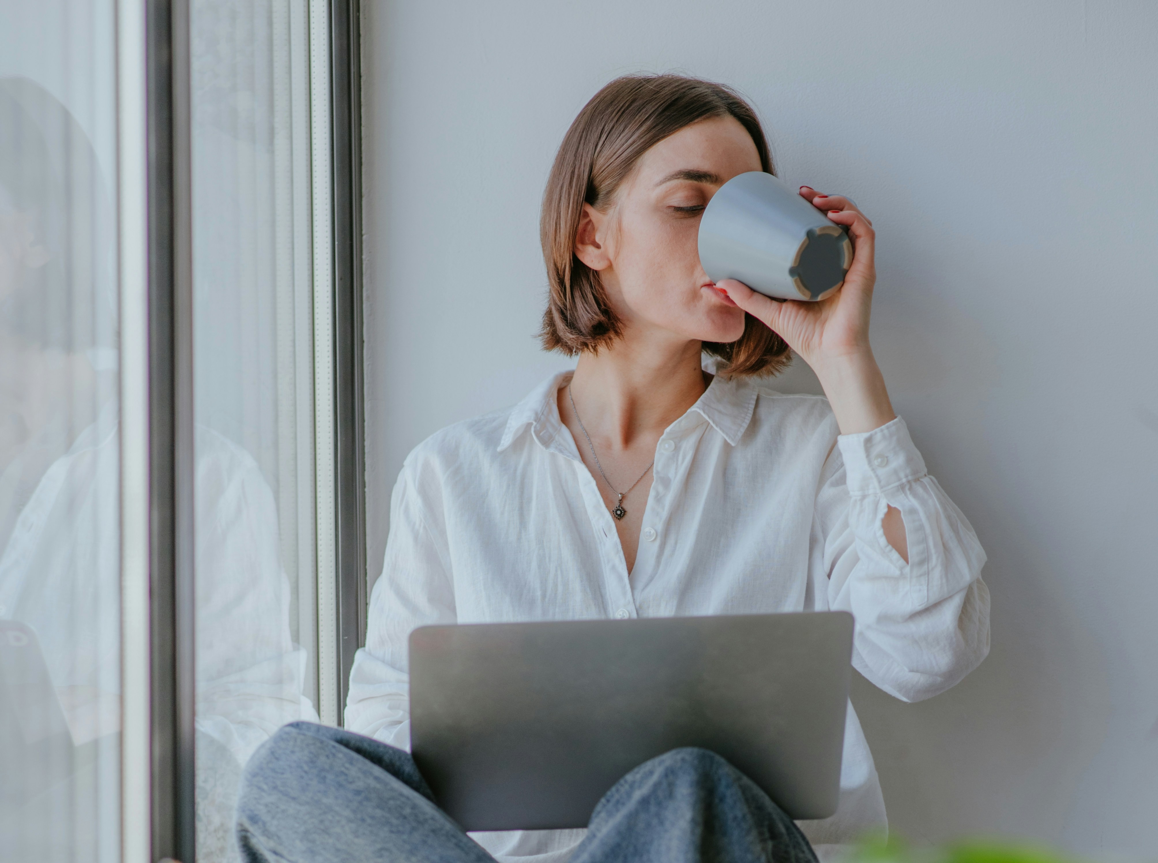 A woman in a white shirt drinks from a mug while sitting with a laptop by a window.