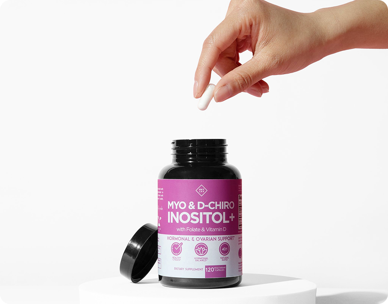 A hand holding a white capsule over an open bottle of Myo & D-Chiro Inositol+ supplements.