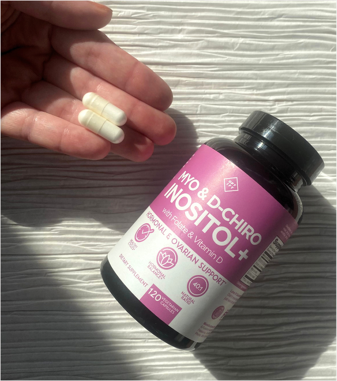 A hand holding two white capsules next to a bottle of Myo & D-Chiro Inositol+ dietary supplement.