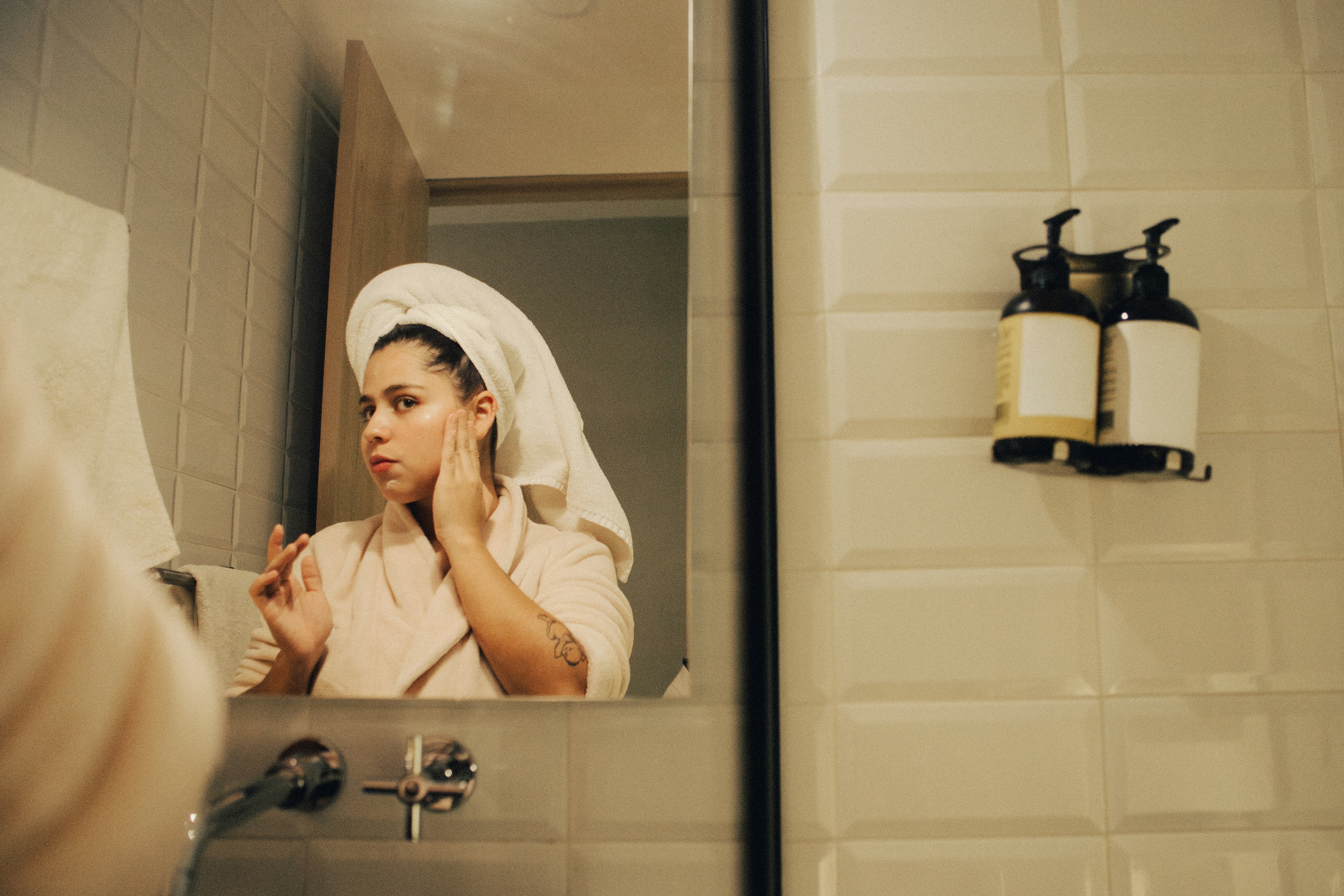 A woman with a towel on her head applies skincare while looking in a bathroom mirror.