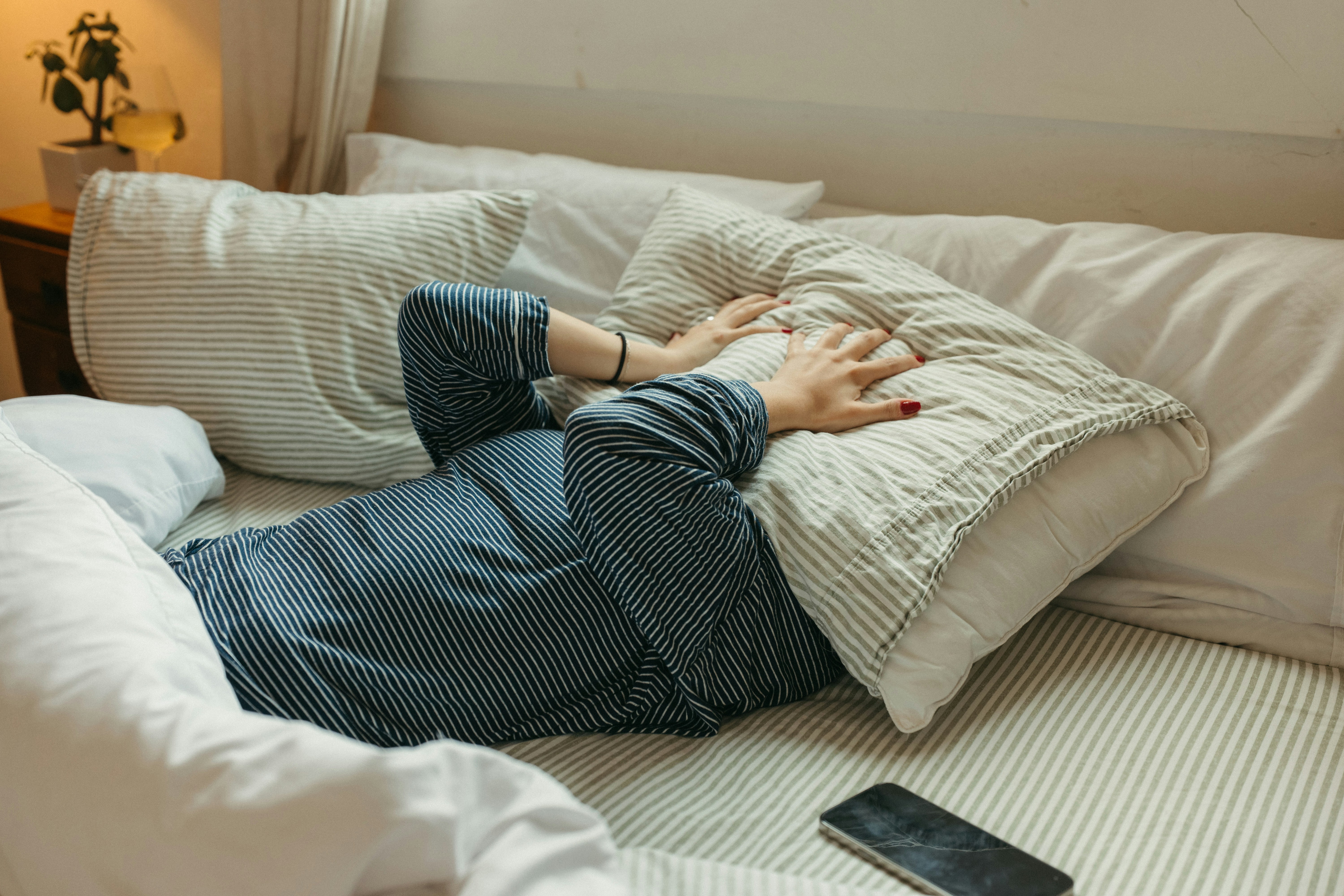 A person in bed covers their head with a pillow, with a cracked smartphone lying next to them.