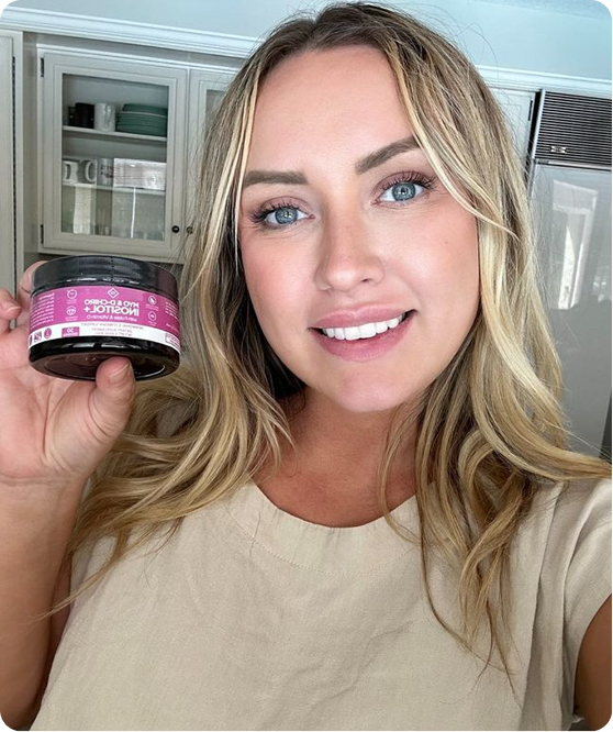A smiling woman with blonde hair holds up a small black jar with a pink label in a kitchen.