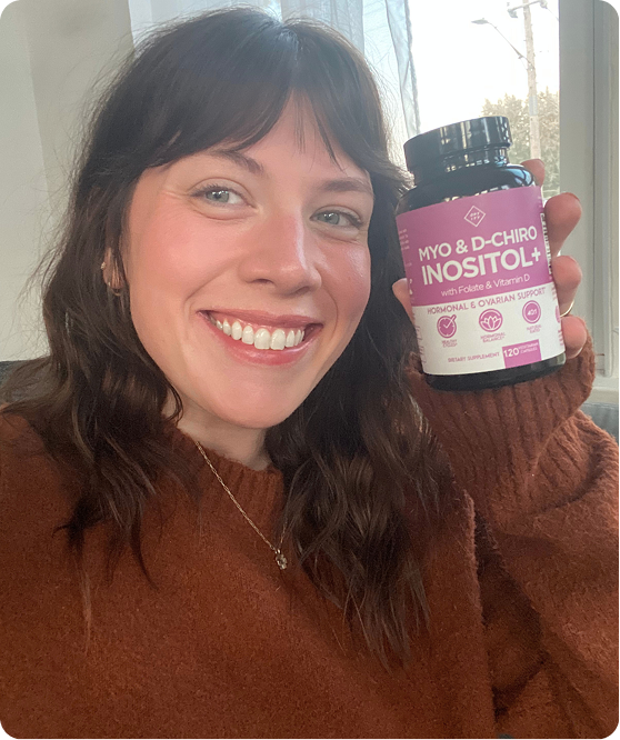 A smiling woman in a brown sweater holds up a bottle of Myo & D-Chiro Inositol+ supplements.