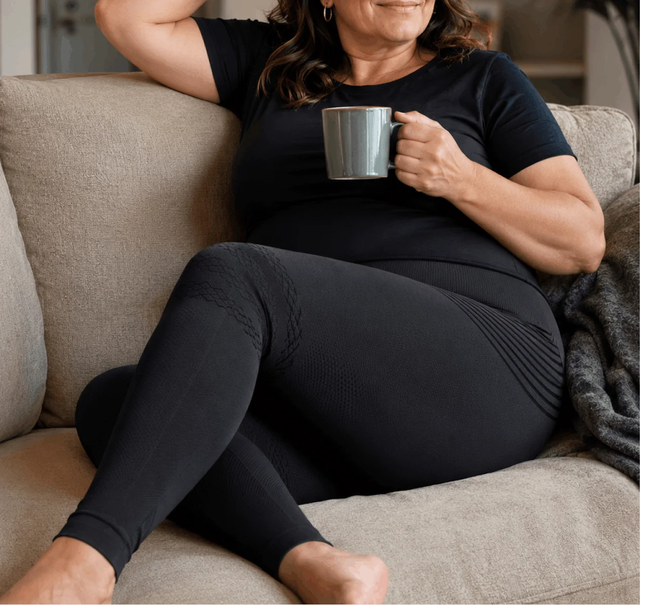 A woman in a black shirt and leggings sits on a couch, holding a grey mug.