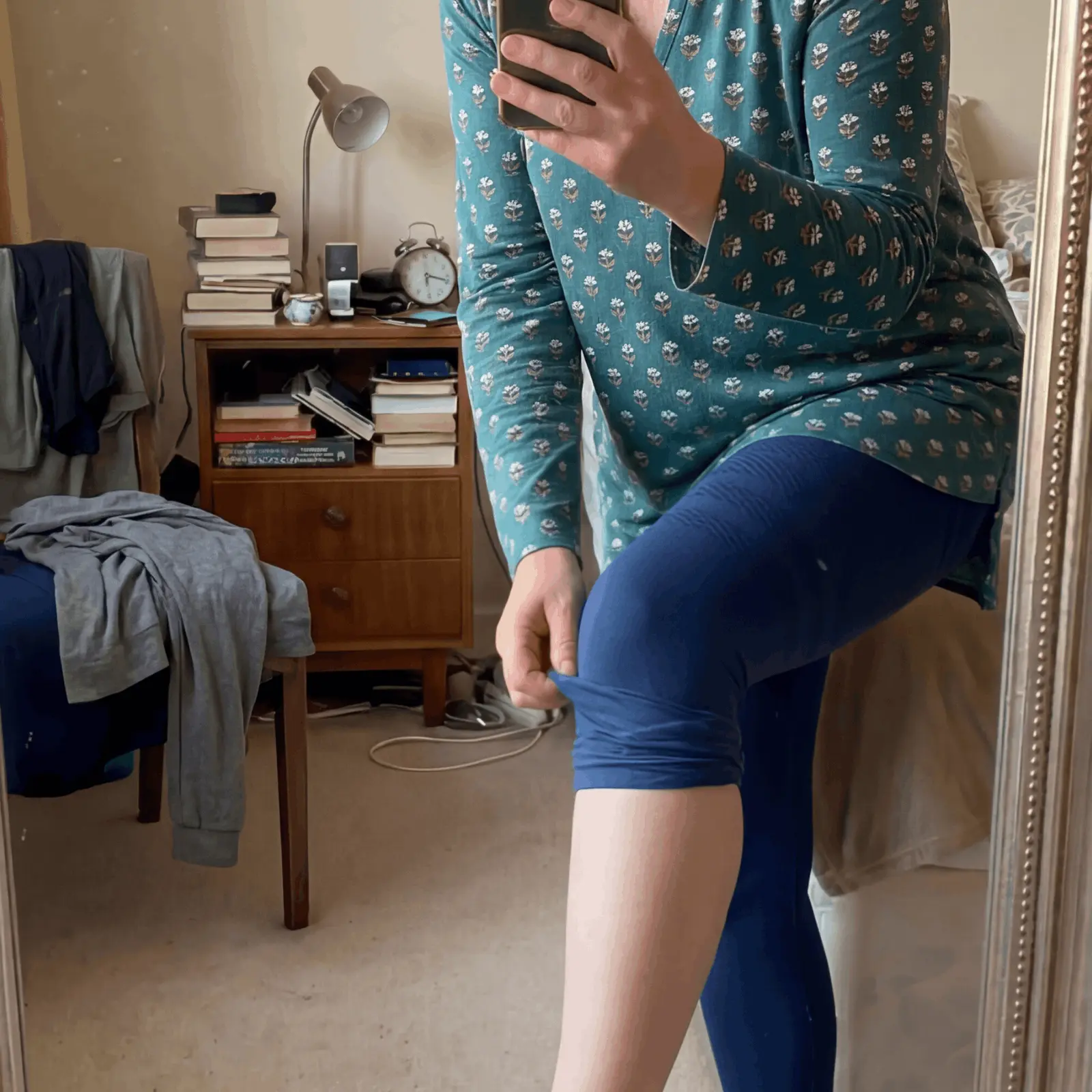 A person in a mirror wearing a floral top and blue leggings, pulling on the legging fabric.