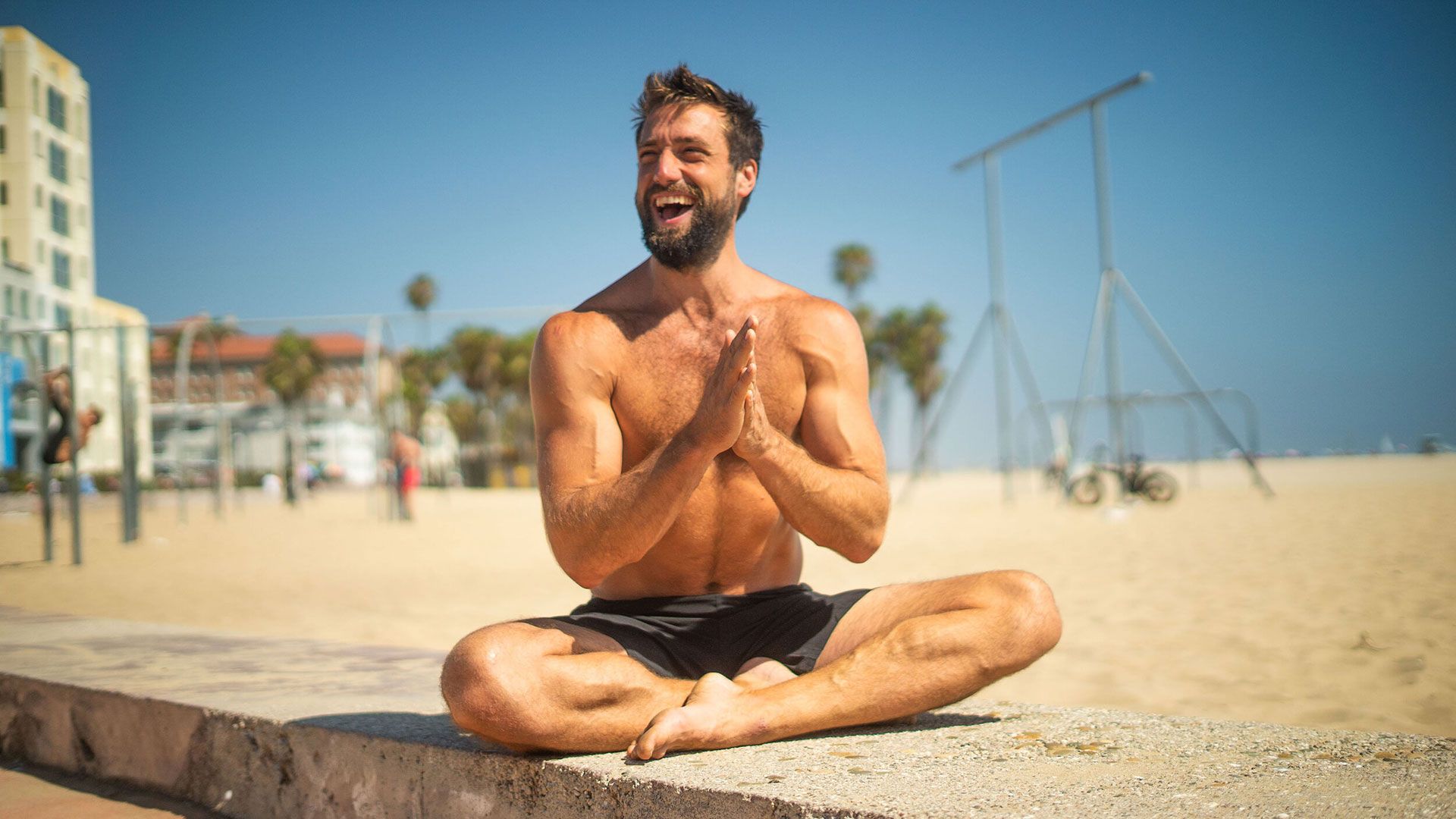 Man sitting cross-legged on a sandy beach, smiling and clapping.