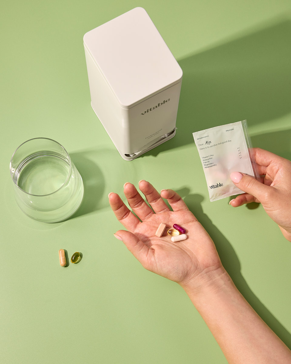A person holds vitamin pills from a personalized packet, with a dispenser and glass of water on a green surface.