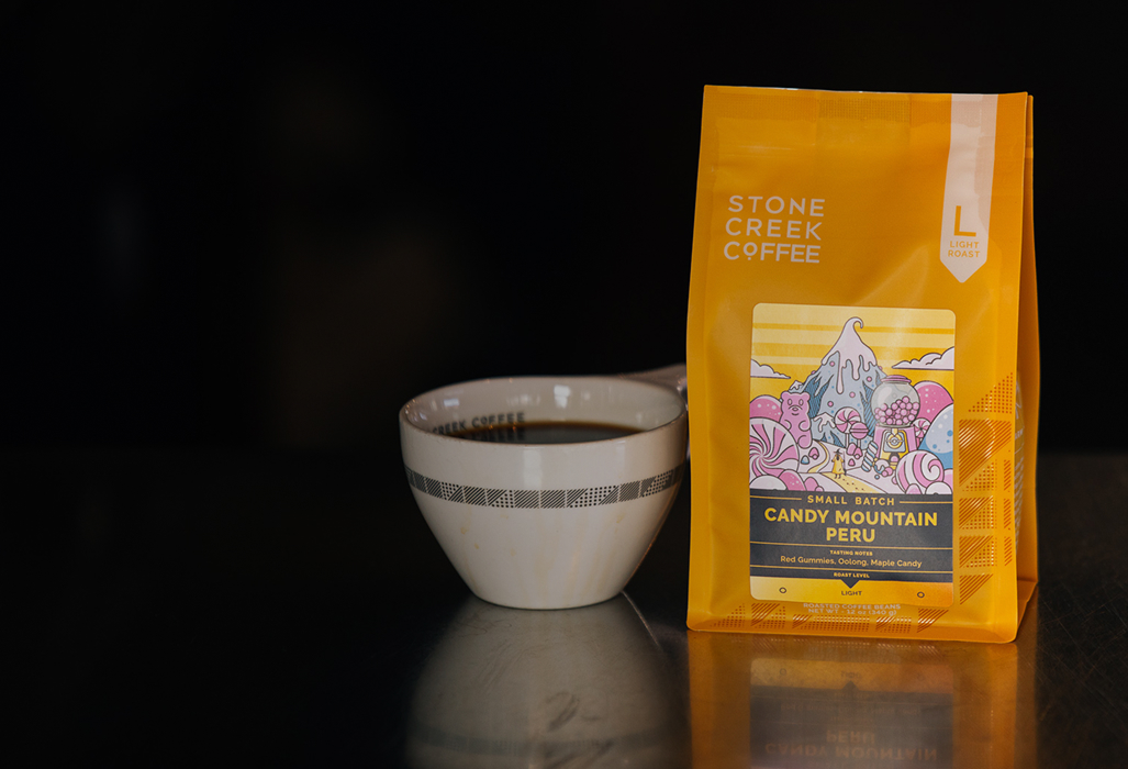 A yellow bag of Stone Creek Coffee sits on a reflective surface next to a white cup of coffee.