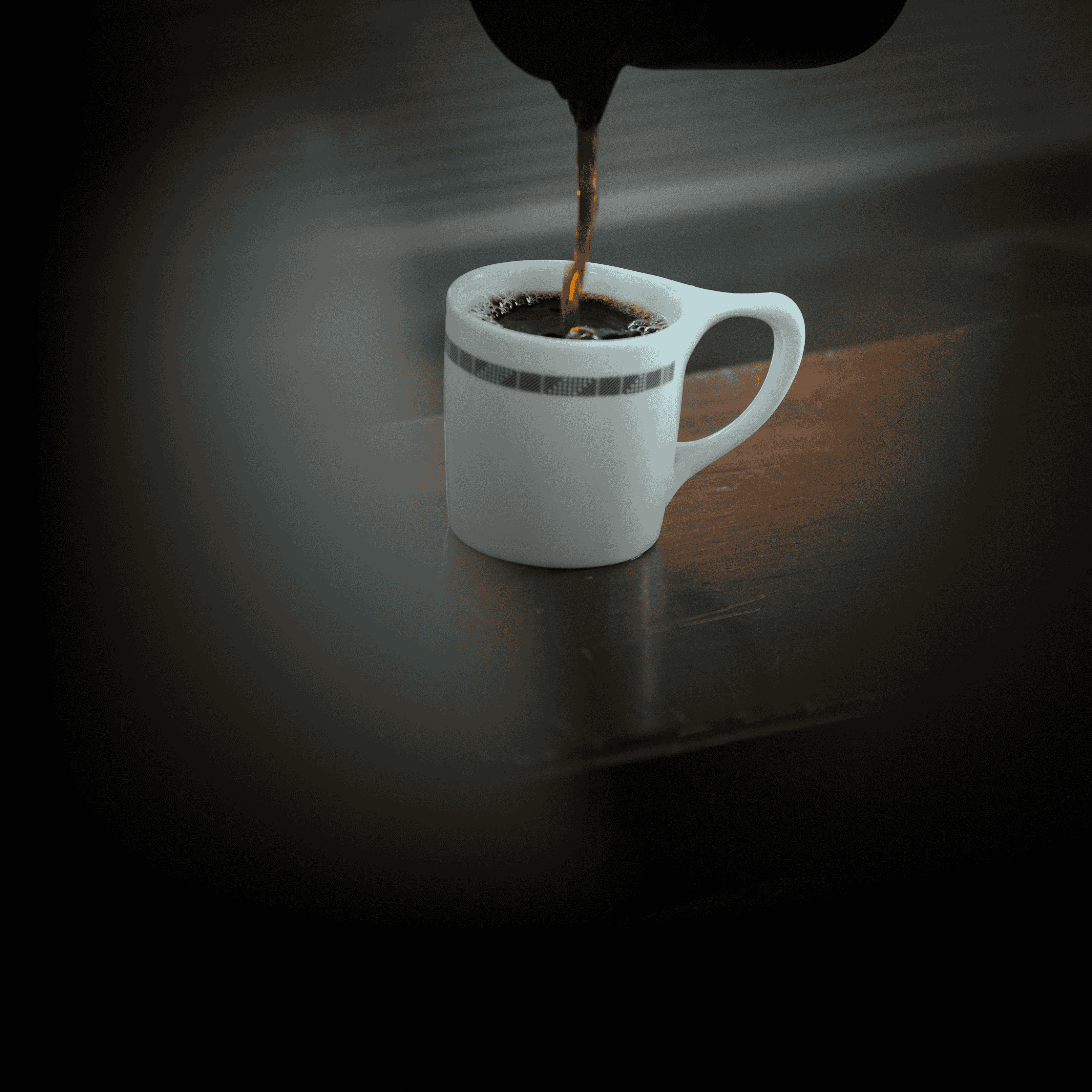 A close-up of dark coffee being poured into a white mug with a patterned rim on a dark surface.