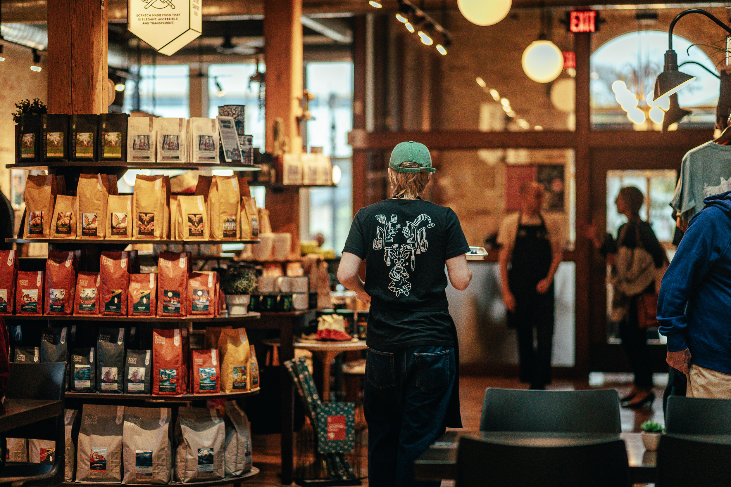 A person seen from behind walks through a warmly lit coffee shop with shelves of coffee beans.