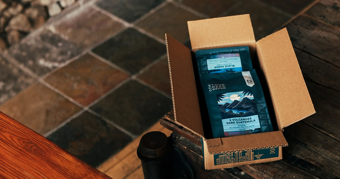 An open cardboard box containing several bags of Stone Creek Coffee on a wooden surface.