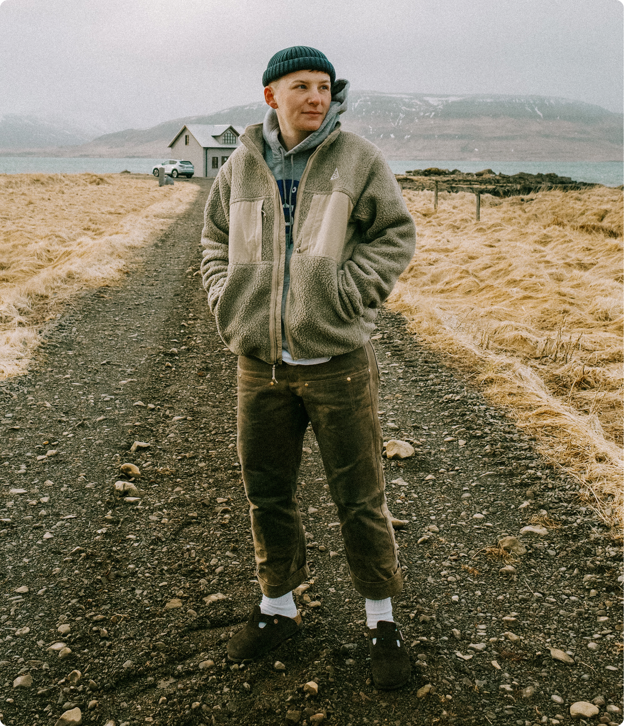 A person in a beanie and fleece jacket stands on a gravel path in a rural landscape.